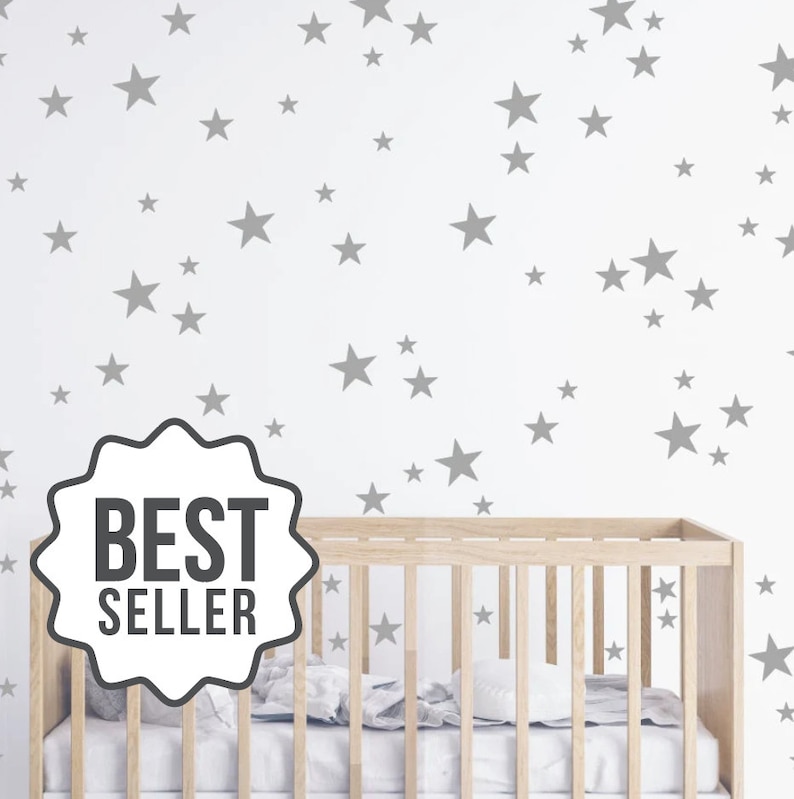 May include: A white crib with a gray star wall decal. The decal features a variety of sizes and shapes of stars. The text "BEST SELLER" is on a white tag.