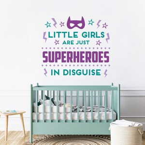 May include: Purple and teal wall decal with the text "Little girls are just superheroes in disguise". The decal features a superhero mask, stars, and lightning bolts. The decal is displayed on a white wall above a light green crib.