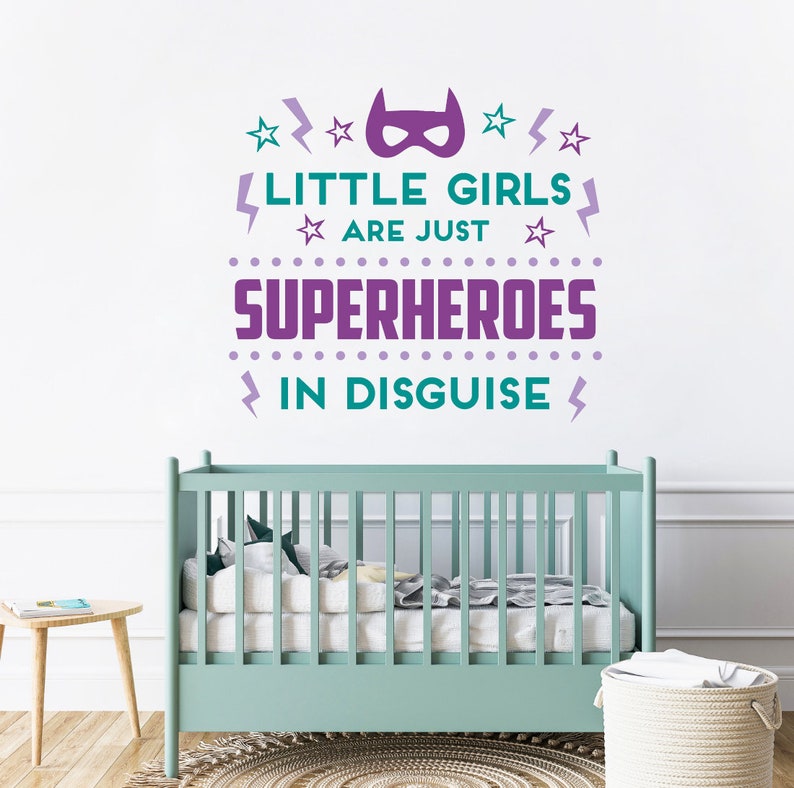 May include: Purple and teal wall decal with the text "Little girls are just superheroes in disguise". The decal features a superhero mask, stars, and lightning bolts. The decal is displayed on a white wall above a light green crib.