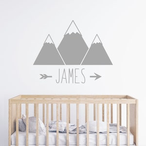 May include: Gray mountain silhouette wall decal with the name "JAMES" and arrows on either side. The decal is above a wooden crib with white bedding.