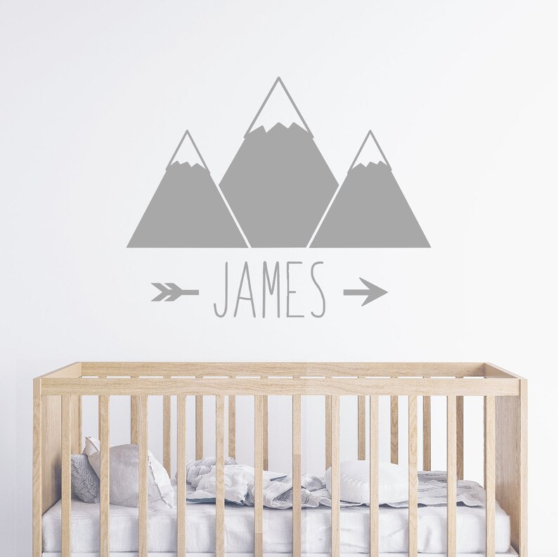 May include: Gray mountain silhouette wall decal with the name "JAMES" and arrows on either side. The decal is above a wooden crib with white bedding.
