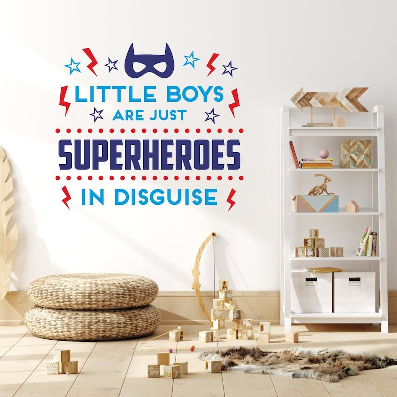 May include: A white wall with a blue and red wall decal that reads "Little Boys Are Just Superheroes In Disguise". The decal features a superhero mask, stars, and lightning bolts. The wall is decorated with a white bookshelf with wooden shelves and a brown wooden arrow.