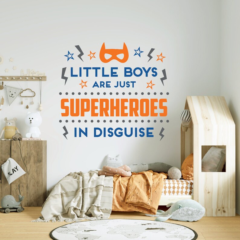 May include: A nursery wall decal with the text "Little boys are just superheroes in disguise". The text is in orange and blue with a superhero mask graphic. The decal is on a white wall in a child's bedroom.