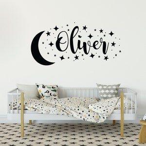 May include: A white cot with black and white star patterned bedding. The wall behind the cot has a black crescent moon and stars with the name "Oliver" written in cursive.