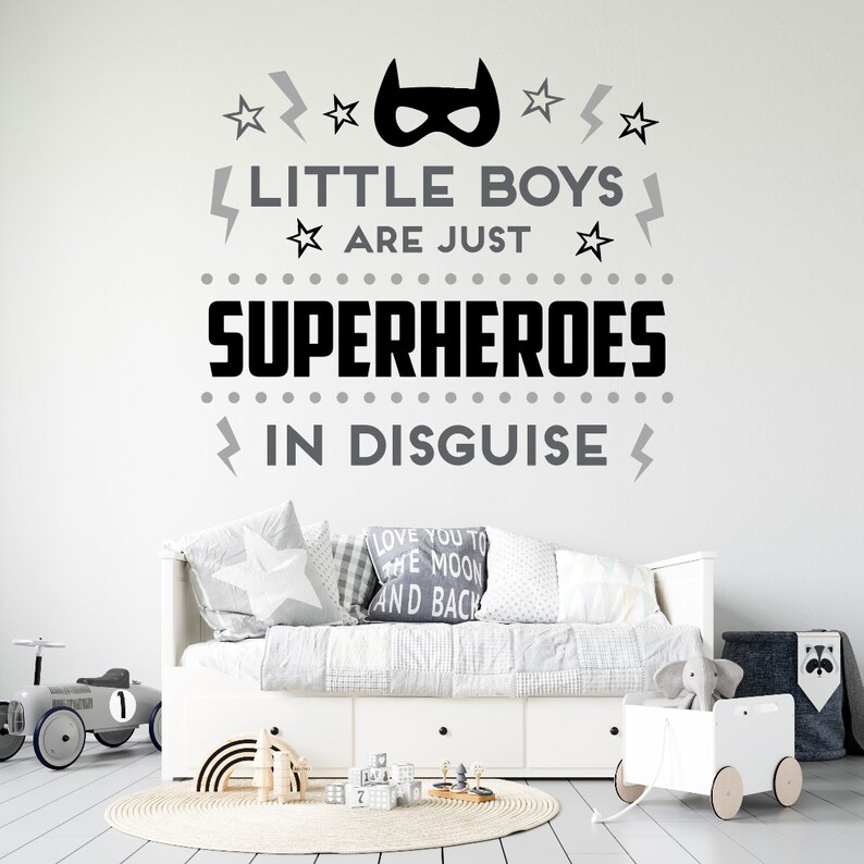 May include: A black and gray wall decal with the text "Little boys are just superheroes in disguise". The decal features a superhero mask, stars, and lightning bolts. The decal is displayed on a white wall above a white daybed with gray and white bedding.