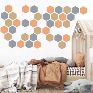 May include: Honeycomb-shaped wall decals in shades of orange, gray, and tan are arranged in a pattern on a white wall. The decals are above a bed with a brown blanket, pillows, and a stuffed whale. The room has a wooden house-shaped bed frame.