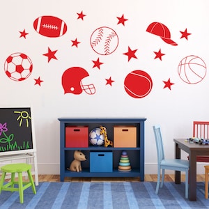 May include: A white wall with red sports-themed decals, including a football, baseball, soccer ball, basketball, a football helmet, and a baseball cap. There are also red stars scattered around the decals. The wall is in a room with a blue bookshelf, a wooden table, and two chairs.