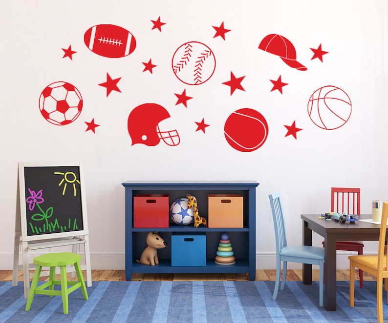 May include: A white wall with red sports-themed decals, including a football, baseball, soccer ball, basketball, a football helmet, and a baseball cap. There are also red stars scattered around the decals. The wall is in a room with a blue bookshelf, a wooden table, and two chairs.