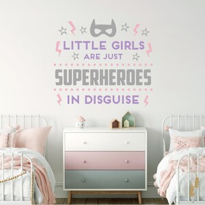 May include: A white wall with a pink and gray vinyl decal that reads "Little girls are just superheroes in disguise". The decal features a superhero mask, stars, and lightning bolts. The decal is displayed above a white dresser with three drawers. The dresser has a pink top drawer and a light blue bottom drawer. There are two white twin beds with pink and white bedding on either side of the dresser.