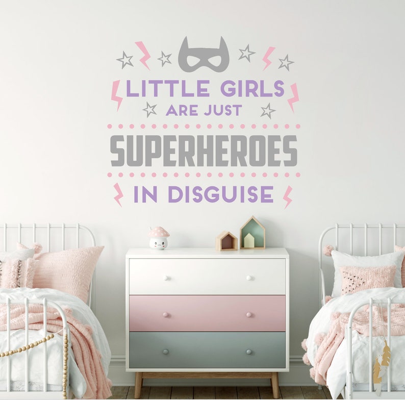May include: A white wall with a pink and gray vinyl decal that reads "Little girls are just superheroes in disguise". The decal features a superhero mask, stars, and lightning bolts. The decal is displayed above a white dresser with three drawers. The dresser has a pink top drawer and a light blue bottom drawer. There are two white twin beds with pink and white bedding on either side of the dresser.