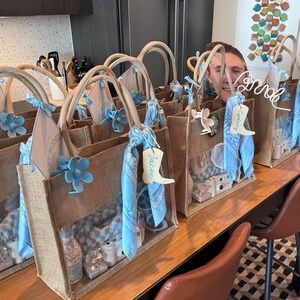 May include: A row of brown burlap gift bags with blue and white checked fabric and blue bandana accents. Each bag has a personalised name tag with a cowboy boot design. The bags are filled with various gifts and are sitting on a wooden table.