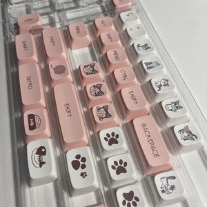 Puede incluir: Teclas de teclado rosa y blancas con diseños de gatos y huellas de patas. Las teclas están dispuestas en una caja de plástico transparente. Las teclas tienen el siguiente texto: MIN, ESC, ALT, SHIFT, MENU, CTRL, SHIFT, BACKSPACE, MENU, SHIFT, SHIFT, INTRO, SHIFT, BLOQ MAYUS, IN.