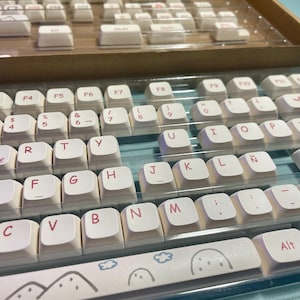 Puede incluir: Teclas blancas de teclado con letras rosas, dispuestas en filas sobre una superficie azul. Las teclas presentan un diseño de montaña y nube en la barra espaciadora.