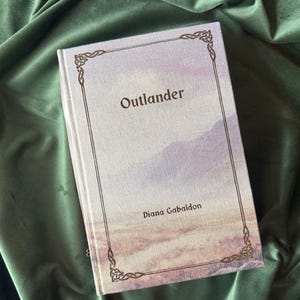 Op de afbeelding: Een hardcover boek getiteld "Outlander" van Diana Gabaldon, met een landschapsontwerp op de omslag. Het boek heeft een decoratieve rand en ligt op een groene fluwelen achtergrond.