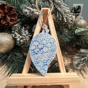 May include: A blue and white ceramic Christmas ornament with a lace-like pattern, hanging from a twine string. The ornament is displayed on a small wooden easel, with a festive background of pine branches, pinecones, and gold ornaments.
