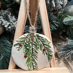 May include: A round, white Christmas ornament with a hand-painted design of green pine needles and red berries. The ornament is suspended by a natural twine cord and a clear bead. The ornament is displayed on a wooden easel, with a backdrop of Christmas greenery.