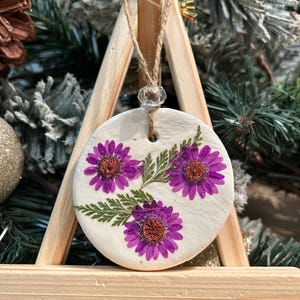 May include: A round, white ornament with three purple flowers and green leaves. The ornament hangs from a twine string with a clear bead. The ornament is displayed on a small wooden easel, with a Christmas tree in the background.