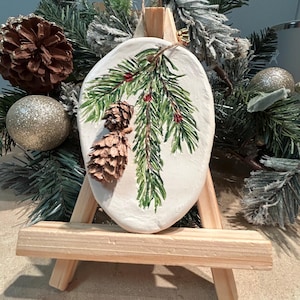 May include: A handmade Christmas ornament featuring a painted evergreen branch with red berries and a pinecone. The ornament is displayed on a small wooden easel, surrounded by festive greenery and gold glitter ornaments.
