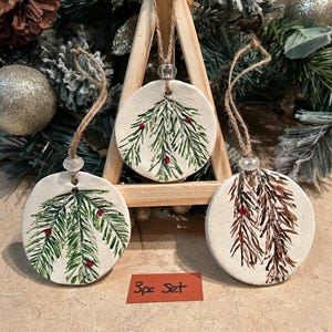 May include: Set of three round, white ceramic Christmas ornaments. Each ornament features a hand-painted evergreen branch design with red berries. The ornaments hang from twine and are displayed on a wooden stand. The text "3pc Set" is on a small brown tag.