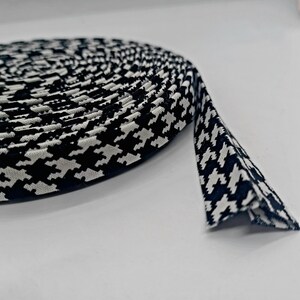 May include: A roll of black and white houndstooth fabric bias tape. The tape is folded over, creating a clean edge. The pattern is a classic, repeating design. The tape is likely used for edging or binding.