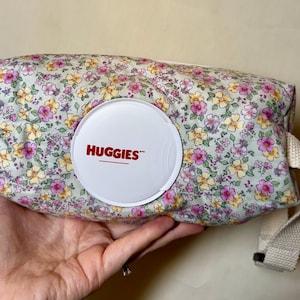 May include: Floral patterned baby wipes container with a white lid featuring the brand name "HUGGIES". The container has a cream-colored strap and is filled with baby wipes. The floral pattern includes pink and yellow flowers.