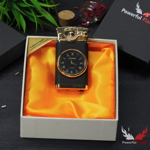 May include: A black and gold lighter with a watch face on the front. The lighter is in a white box with a yellow satin lining. The box has the words "LIGHTER CLASSICAL FASHIONABLE" printed on the side. The box is sitting on a black surface.