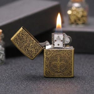May include: A brass-toned metal lighter with an open lid and a visible flame. The exterior features an intricate embossed pattern and a central circular emblem containing the letters C S S M L N D S M D, surrounded by a decorative border design.