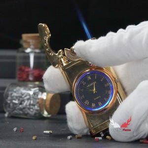 May include: A gold-tone lighter with a built-in watch face. The watch face has black Roman numeral hour markers and a blue background. The lighter is lit and has a blue flame.