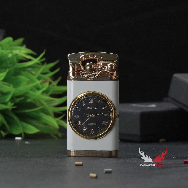 May include: A white lighter with a gold-tone finish and a built-in analogue clock face. The clock face has black Roman numeral hour markers and a black background. The lighter has a gold-tone lever on the top.