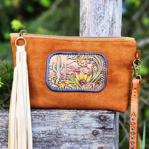 May include: A brown leather clutch with a decorative desert scene, featuring a cactus and flowers, is displayed. The clutch has a long cream-colored tassel and a matching wrist strap. The design is reminiscent of the American Southwest.