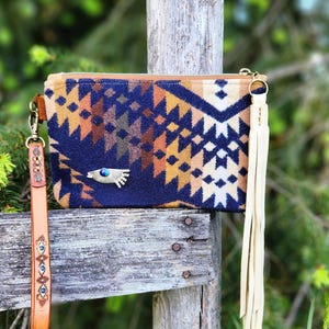 May include: A rectangular clutch with a geometric pattern in navy blue, tan, and cream. It has a leather wrist strap and a cream-colored tassel. A silver-toned charm with a turquoise stone is on the front. The clutch is displayed on a wooden fence.