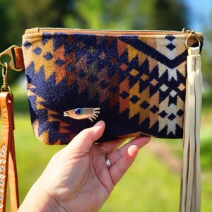 Puede incluir: Un bolso de mano estampado con una correa de mu&ntilde;eca de cuero marr&oacute;n y una borla color crema. El bolso de mano presenta un dise&ntilde;o geom&eacute;trico en colores azul marino, marr&oacute;n y crema. Se adjunta un broche dorado en forma de pez con una gema azul.