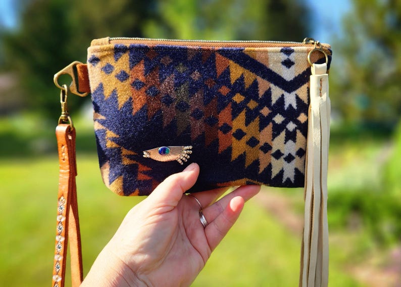 Puede incluir: Un bolso de mano estampado con una correa de mu&ntilde;eca de cuero marr&oacute;n y una borla color crema. El bolso de mano presenta un dise&ntilde;o geom&eacute;trico en colores azul marino, marr&oacute;n y crema. Se adjunta un broche dorado en forma de pez con una gema azul.