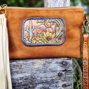 Puede incluir: Un bolso de mano de ante marr&oacute;n con un parche decorativo de cuero que representa una escena des&eacute;rtica con un cactus y un girasol. Tiene una borla color crema y una correa de mu&ntilde;eca de cuero marr&oacute;n con tachuelas decorativas.