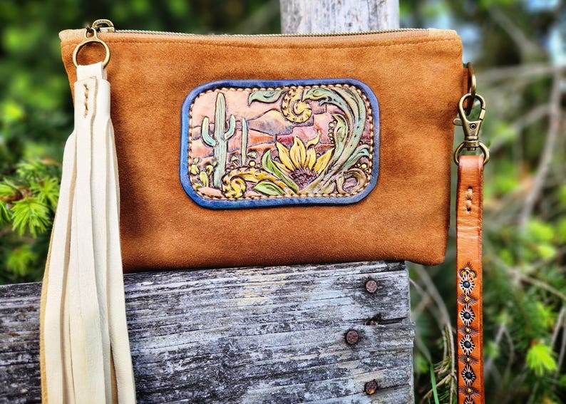 Puede incluir: Un bolso de mano de ante marr&oacute;n con un parche decorativo de cuero que representa una escena des&eacute;rtica con un cactus y un girasol. Tiene una borla color crema y una correa de mu&ntilde;eca de cuero marr&oacute;n con tachuelas decorativas.