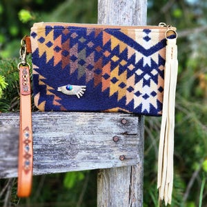 Puede incluir: Bolso de mano estampado con cierre de cremallera, con un dise&ntilde;o geom&eacute;trico en azul marino, marr&oacute;n y naranja. Tiene una correa de mu&ntilde;eca de cuero y una borla color crema. Un adorno en forma de ojo est&aacute; en la parte delantera.