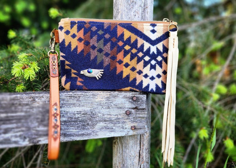 Puede incluir: Bolso de mano estampado con cierre de cremallera, con un dise&ntilde;o geom&eacute;trico en azul marino, marr&oacute;n y naranja. Tiene una correa de mu&ntilde;eca de cuero y una borla color crema. Un adorno en forma de ojo est&aacute; en la parte delantera.