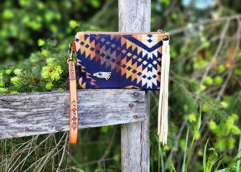 May include: A rectangular clutch with a geometric pattern in navy blue, tan, and cream. It has a leather wrist strap and a cream-colored tassel. A silver-toned charm with a turquoise stone is on the front. The clutch is displayed on a wooden fence.