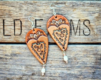 May include: Hand-tooled leather earrings with a heart design. The earrings are a light brown color with floral and turquoise accents. Each earring has a small white bead at the bottom and silver-colored ear hooks. The earrings are displayed on a wooden surface.