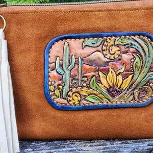 Puede incluir: Bolso de mano de ante marr&oacute;n con un parche decorativo de cuero que representa un paisaje des&eacute;rtico con cactus y un girasol. El parche tiene un borde azul y est&aacute; hecho a mano con detalles intrincados. Una borla crema cuelga de la cremallera.