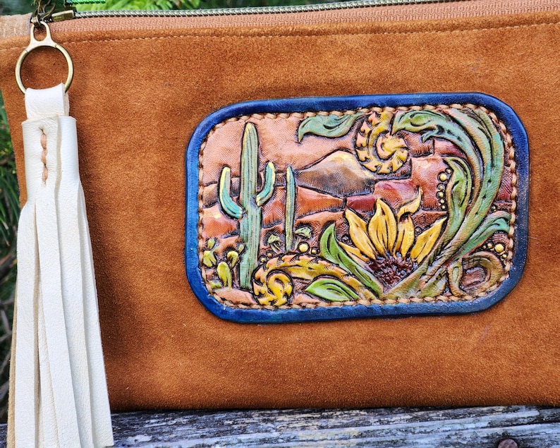 Puede incluir: Bolso de mano de ante marr&oacute;n con un parche decorativo de cuero que representa un paisaje des&eacute;rtico con cactus y un girasol. El parche tiene un borde azul y est&aacute; hecho a mano con detalles intrincados. Una borla crema cuelga de la cremallera.