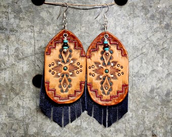 May include: A pair of tan leather earrings featuring a stamped geometric pattern with red and turquoise accents. Each earring has a silver-tone hook, a small turquoise and dark bead detail, and a long dark blue suede fringe at the bottom.