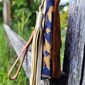 Puede incluir: Una peque&ntilde;a bolsa de cuero marr&oacute;n con un patr&oacute;n geom&eacute;trico azul y beige. La bolsa tiene flecos de cuero color crema y una correa de cuero marr&oacute;n. La bolsa est&aacute; colgada de un poste de valla de madera.