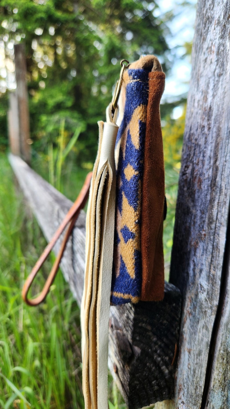 Puede incluir: Una peque&ntilde;a bolsa de cuero marr&oacute;n con un patr&oacute;n geom&eacute;trico azul y beige. La bolsa tiene flecos de cuero color crema y una correa de cuero marr&oacute;n. La bolsa est&aacute; colgada de un poste de valla de madera.