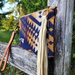 Puede incluir: Un bolso de mano estampado con una correa de mu&ntilde;eca de cuero y una borla color crema. El bolso de mano presenta un dise&ntilde;o geom&eacute;trico en azul marino, naranja y marr&oacute;n. La correa de mu&ntilde;eca es marr&oacute;n con tachuelas met&aacute;licas decorativas. La cremallera es dorada.