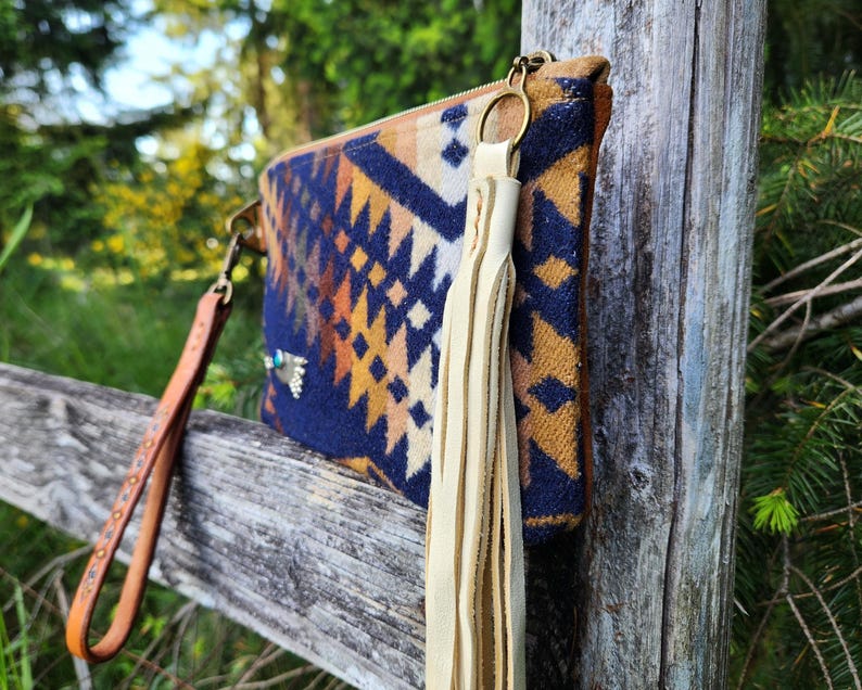 Puede incluir: Un bolso de mano estampado con una correa de mu&ntilde;eca de cuero y una borla color crema. El bolso de mano presenta un dise&ntilde;o geom&eacute;trico en azul marino, naranja y marr&oacute;n. La correa de mu&ntilde;eca es marr&oacute;n con tachuelas met&aacute;licas decorativas. La cremallera es dorada.