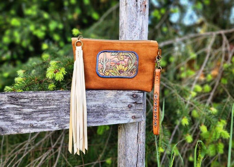 May include: A brown leather clutch with a decorative desert scene, featuring a cactus and flowers, is displayed. The clutch has a long cream-colored tassel and a matching wrist strap. The design is reminiscent of the American Southwest.