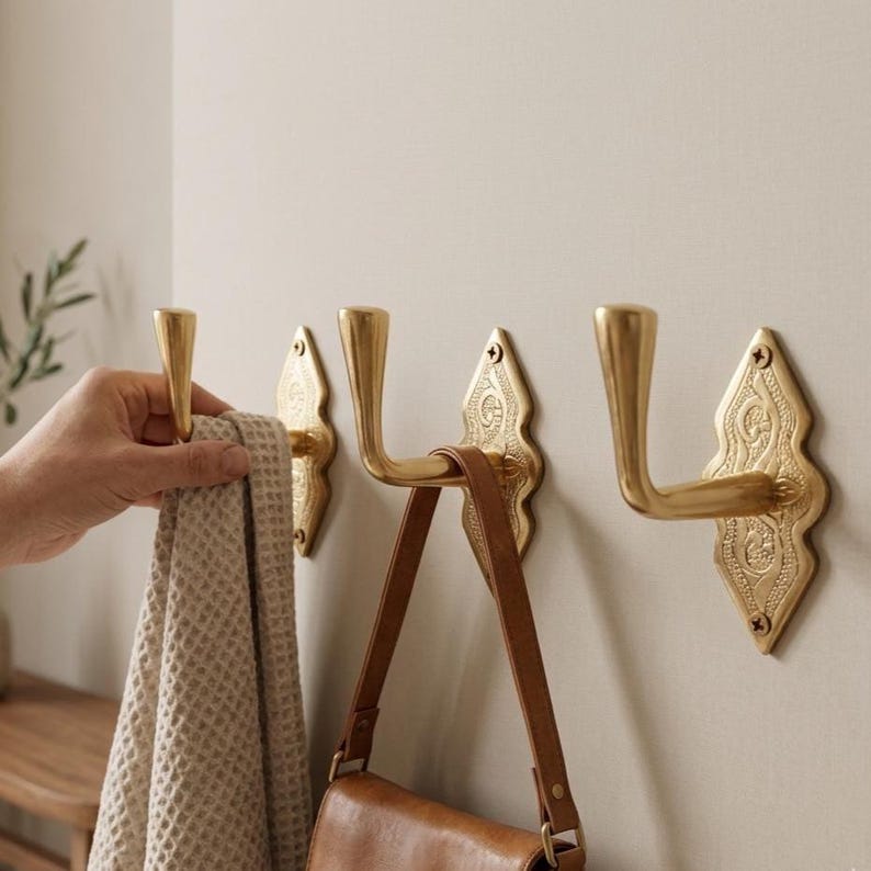 May include: Three gold-colored metal wall hooks with a diamond-shaped backplate featuring an embossed pattern. Each hook has a curved arm and is secured with two screws. A beige waffle-textured towel hangs from the leftmost hook.