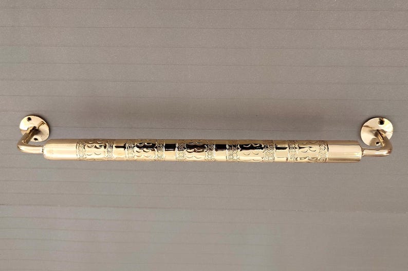 K&ouml;nnte beinhalten: Eine goldfarbene Metall-Handtuchhalterung mit einem verzierten Design. Die Stange hat abgerundete Enden und ist an einer Wand montiert.