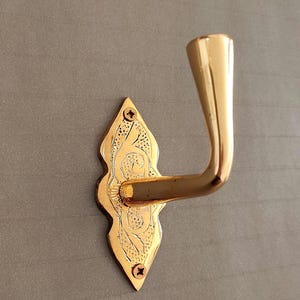May include: A polished gold-tone metal wall hook featuring a decorative, elongated diamond-shaped backplate with intricate etched detailing. The hook is secured to the wall with two visible screws at the top and bottom of the backplate.